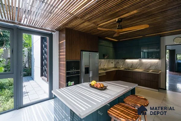 A kitchen with marble counter tops and wooden cabinetry, along with an island in the middle of the middle of the island with stairs leading to shop & more windows overlooking the ocean in the background at night time